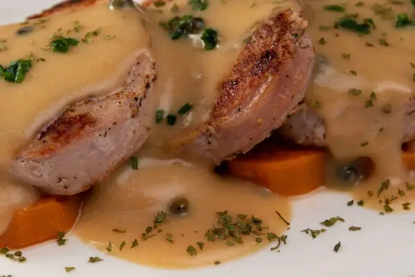 Tranches de gigot d'agneau nappées d'une sauce onctueuse au fond de veau, parsemées de persil et de câpres, sur un lit de carottes.