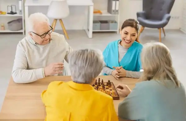 Un gruppo di anziani gioca a scacchi, un'attività che promuove la stimolazione cognitiva. Un'assistente sorride osservando.