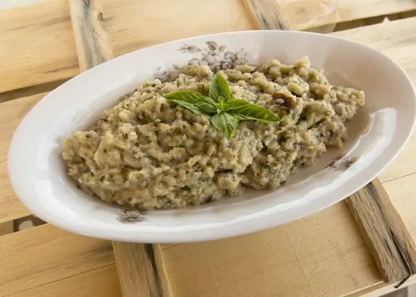 Hummus di melanzane cremoso, guarnito con basilico fresco, servito in un piatto ovale su un tavolo di legno grezzo.