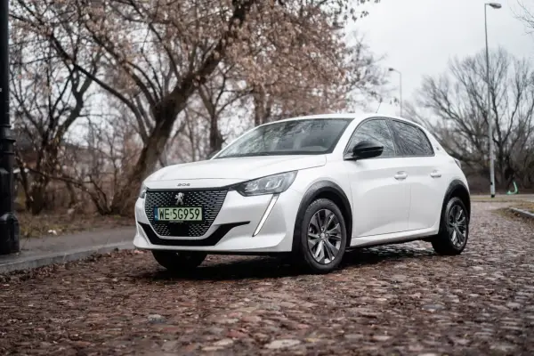 Biały Peugeot e-208 na brukowanej ulicy, z drzewami w tle.