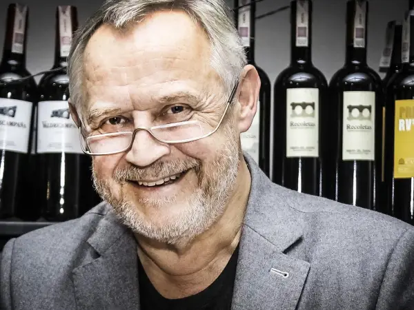 Marek Kondrat, z uśmiechem i okularami, na tle butelek wina. Jego majątek to nie tylko sukcesy zawodowe, ale i pasja do trunków.