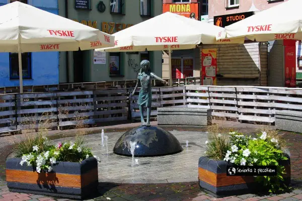 Fontanna z dziewczynką na kuli, otoczona donicami z kwiatami i parasolami. Urokliwe lubliniec atrakcje, idealne na spacer.