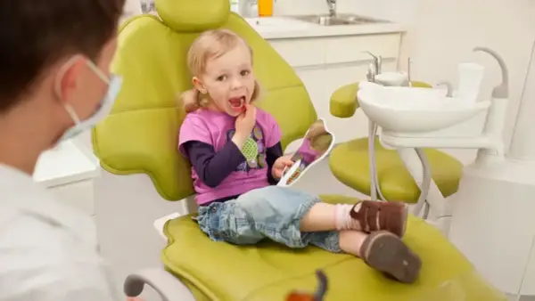 Mała dziewczynka z lusterkiem sprawdza swoje zdrowe zęby mleczne podczas wizyty u dentysty.
