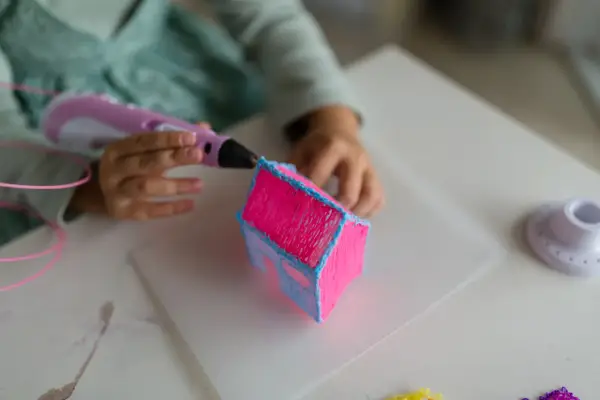 Dziecko tworzy różowy domek z niebieskimi detalami za pomocą różowego długopisu 3D.