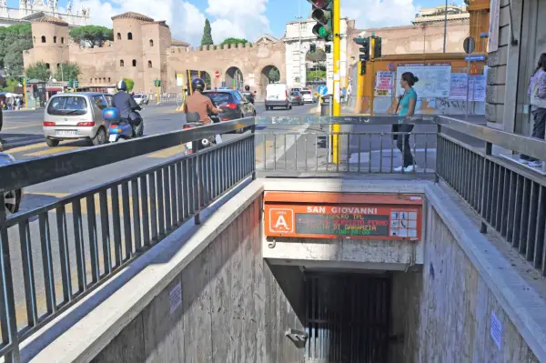 Ingresso metro San Giovanni, collegamento metro A. Traffico intenso con auto e scooter, sullo sfondo le antiche mura romane.