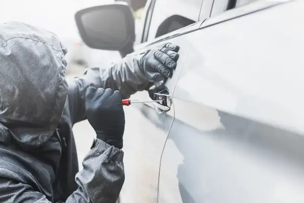 Osoba w kapturze i rękawiczkach próbuje otworzyć auto śrubokrętem. Sen o kradzieży auta może symbolizować utratę kontroli.