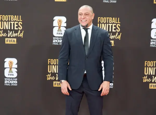 Roberto Carlos żyje i wita fanów na czerwonym dywanie, ubrany w elegancki garnitur. Uśmiecha się szeroko, a w tle widać logo FIFA i napisy związane z Mistrzostwami Świata.