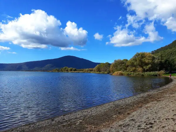 Spiaggia ghiaiosa e acque calme del lago di Vico, con montagne verdi e cielo azzurro punteggiato di nuvole bianche.