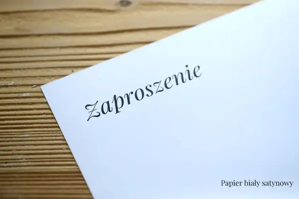 Eleganckie zaproszenie na ślub na białym, satynowym papierze, leżące na drewnianym tle.