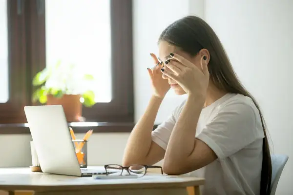 Zmęczona kobieta zamyka oczy, czując brak zbieżności objawy. Przed nią laptop i okulary.
