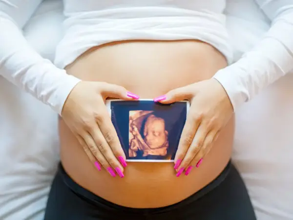 Ręce z różowymi paznokciami obejmują brzuch, na którym widnieje zdjęcie z 8 tygodnia ciąży usg 3d.
