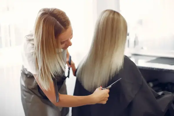 Peluquera con tijeras realiza un corte de cabello en v a una clienta con cabello rubio largo.