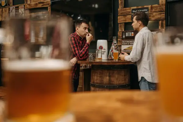 Dwóch mężczyzn rozmawia przy barze, na którym stoi kufel i szklanka piwa. Dyskutują, ile schodzi jedno piwo, podczas gdy w tle widać drewniane dekoracje i plakaty.