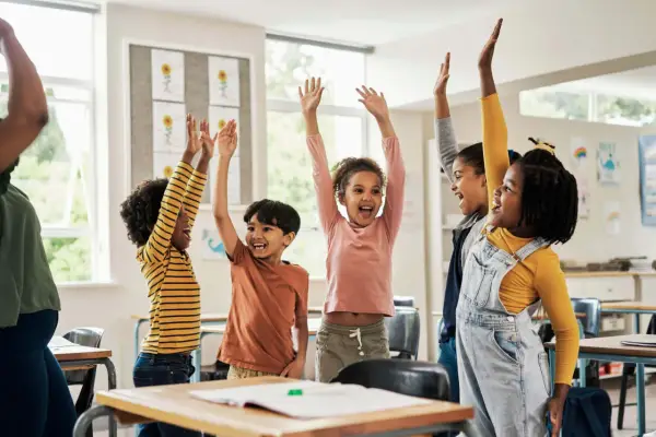 Fröhliche Kinder mit erhobenen Armen in einem Klassenzimmer. Sie nehmen begeistert an Kennenlernspielen für die Grundschule teil.