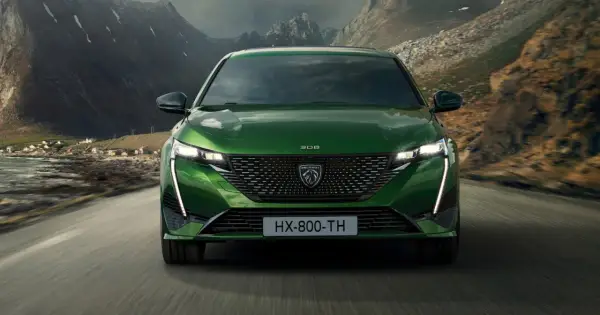 Zielony Peugeot 308 T7 jedzie drogą w malowniczym, górskim krajobrazie z widokiem na małą wioskę nad wodą.