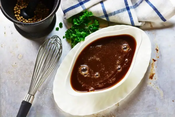 Sauce à la viande fumante dans un saucier, avec un fouet, du persil et un mortier rempli de grains de poivre.
