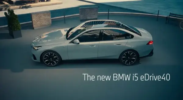 Jasnoszary sedan BMW i5 eDrive40 stoi na zewnątrz, z widokiem na basen i ławkę.
