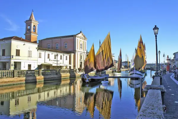Traditional sailboats with colorful sails line a canal in Cesenatico, Italy, with buildings and a church reflected in the water.