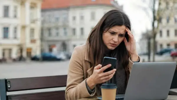 Zmartwiona kobieta czyta na telefonie coś, co wygląda na zniesławienie. Trzyma się za głowę, siedząc przy stoliku z laptopem i kawą.