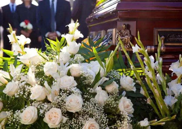 Elegancki wieniec pogrzebowy z białych róż i lilii, ozdobiony zielenią, przy trumnie. Cena 200 zł.