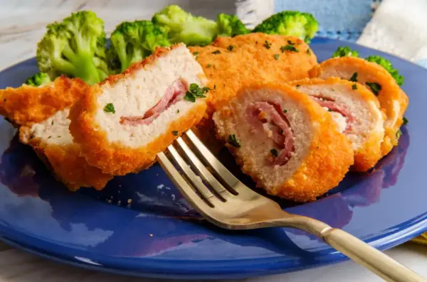 Kotlet po szwajcarsku, panierowany, z szynką w środku, podany z brokułami na niebieskim talerzu. Złoty widelec czeka na pierwszy kęs.