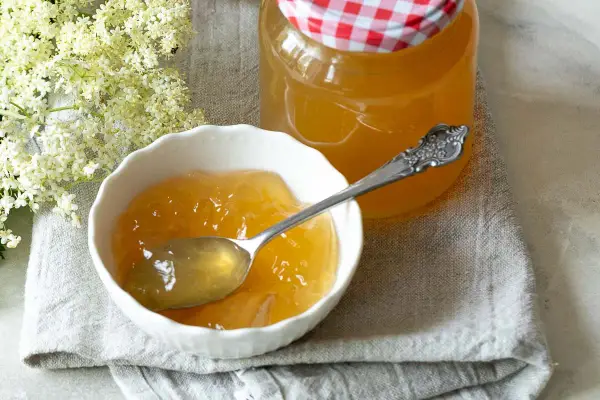 Ein Löffel voll goldgelbem Holunder Apfel Gelee in einer Schale, daneben ein Glas mit dem Gelee und Holunderblüten.