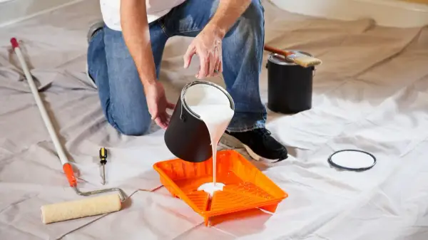 Uomo versa vernice bianca in una vaschetta arancione, pronto per pitturare soffitto con pennello. Attrezzi da pittura a portata di mano.