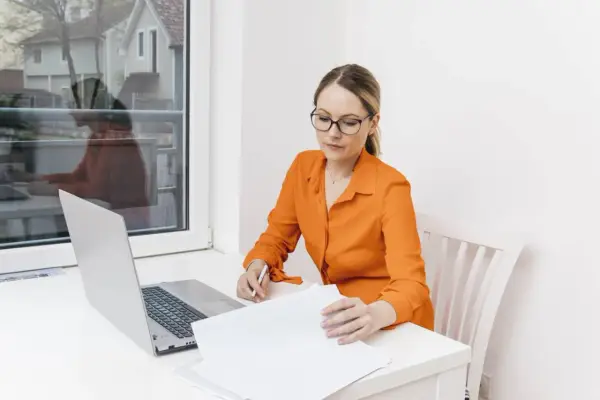 Kobieta w okularach i pomarańczowej bluzce analizuje dokumenty przy laptopie. Dowiaduje się, jak zostać notariuszem.
