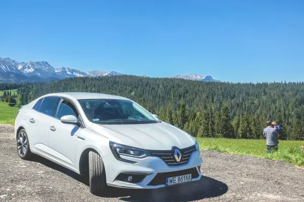Biały Renault Megane Sedan zaparkowany na szutrowej drodze z widokiem na góry i las. Na trawie stoi mężczyzna z dzieckiem.