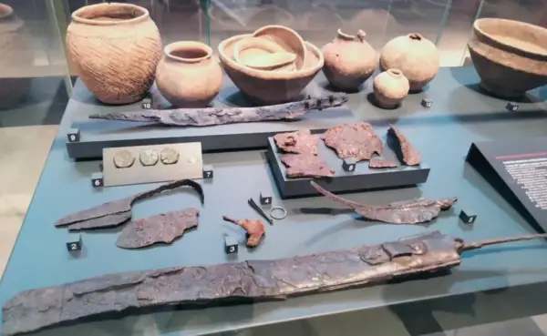 Reperti archeologici, tra cui vasi, monete e armi, simili a quelli della patera di Parabiago, esposti in un museo.