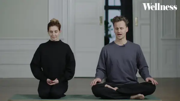 Två personer sitter på en yogamatta och mediterar. En kvinna i svart tröja och en man i grå tröja. Ta ett ögonblick att stanna upp.