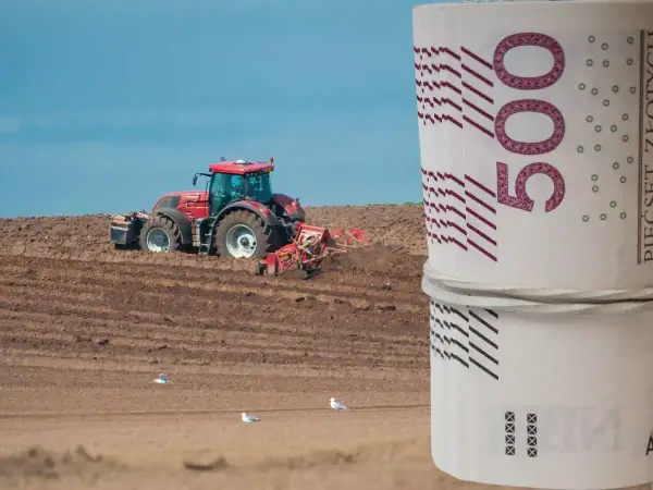 Czerwony traktor orze pole, a na pierwszym planie zwinięte banknoty 500 zł. Zastanawiasz się, ile kosztuje hektar ziemi budowlanej?