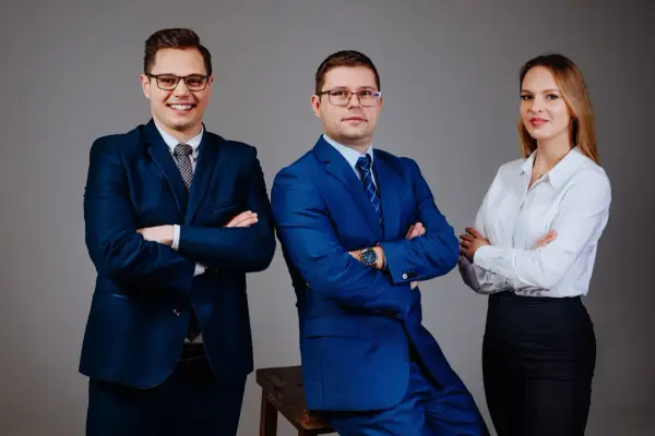 Trzy osoby w garniturach i białej koszuli, stojące na szarym tle, z rękami skrzyżowanymi na piersi.