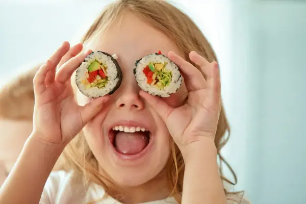 Uśmiechnięta dziewczynka trzyma kawałki sushi dla dzieci przed oczami, udając, że ma rolki zamiast oczu.