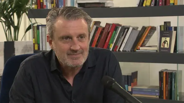 Uomo con barba grigia sorride, microfono davanti. Alle sue spalle scaffali pieni di libri, tra cui "Le ossa parlano trama".