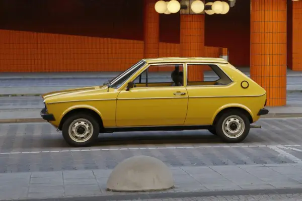 Żółty Audi 50, klasyk z lat 70., parkuje na tle pomarańczowej, kafelkowej architektury.