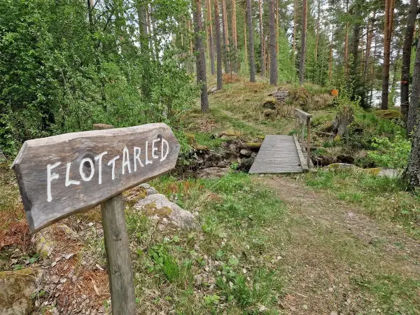 Träskylt med texten "Flottarleden Munkedal" pekar mot en liten träbro över en bäck i en skogsmiljö.
