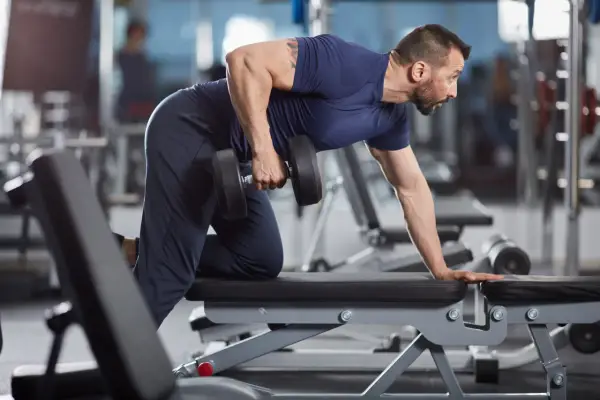 Man utför enarms hantelrodd på ett gym. Han stöttar sig mot en bänk med ena handen och drar en hantel mot bröstet med den andra.