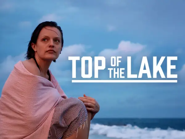 Kobieta owinięta w różowy ręcznik siedzi na plaży, patrząc w dal. Na niebie widnieje napis "TOP OF THE LAKE".