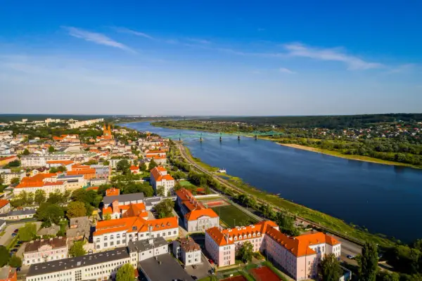 Widok z lotu ptaka na miasto nad Wisłą, z zielonym mostem i zabudowaniami w słoneczny dzień.