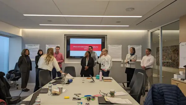 A team coach leads a meeting in a conference room, with a screen displaying "What will make today successful for you?".