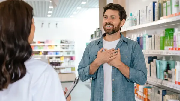 Mężczyzna z dłońmi na piersi rozmawia z farmaceutką, zastanawiając się, ile zarabia farmaceuta.