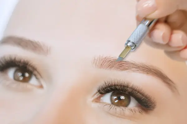 Main d'une esthéticienne réalisant un microblading poil à poil sur un sourcil.