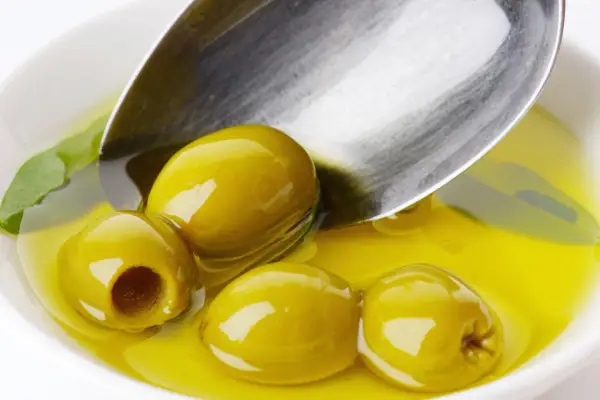 Kilka zielonych oliwek w złocistej oliwie, łyżka zanurzona w płynie. Oliwa z oliwek kcal - zdrowie i smak.