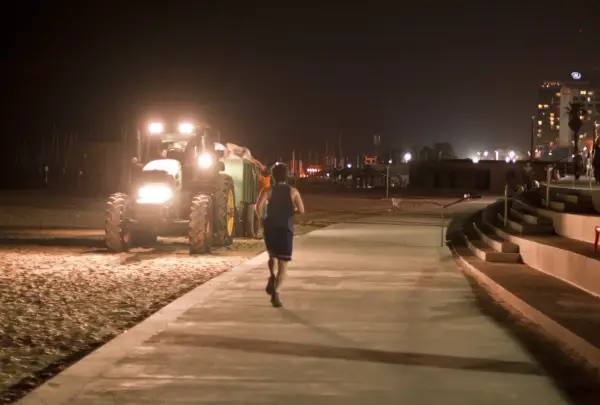 Mężczyzna uprawia bieganie w nocy na promenadzie, obok pracującego traktora na plaży.