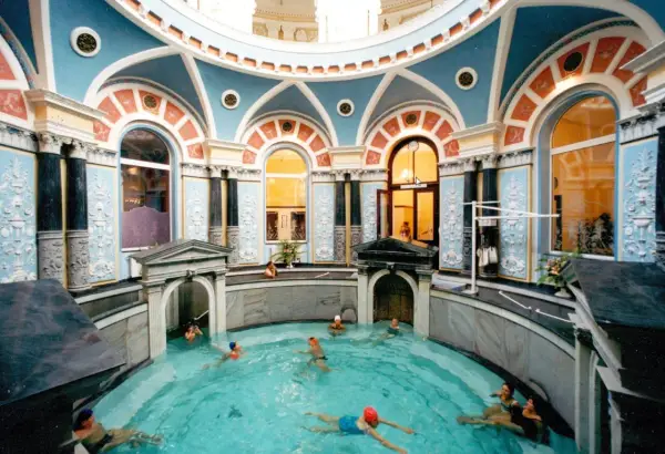 Wnętrze z basenem, gdzie ludzie relaksują się w gorącym źródle. Architektura jest bogato zdobiona, z niebieskimi i pomarańczowymi detalami.