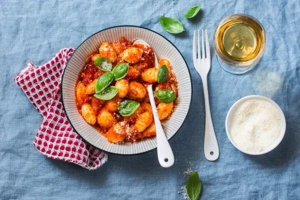 Pyszne kluski gnocchi w sosie pomidorowym, udekorowane bazylią. Obok kieliszek białego wina i miseczka parmezanu.