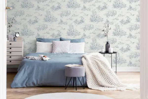 Chambre paisible avec un papier peint au motif jouy bleu et blanc, un lit bleu, des coussins blancs et une couverture en maille.