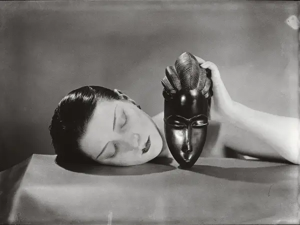 Foto Man Ray: volto femminile addormentato accanto a una maschera africana tenuta da una mano.
