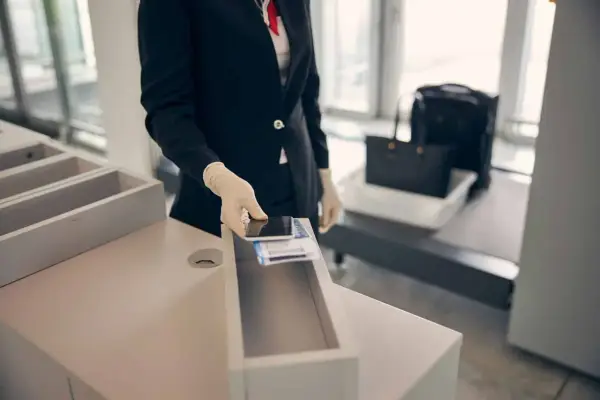 Pracownik lotniska w rękawiczkach kładzie telefon i karty pokładowe do pojemnika. W tle widać bagaż na taśmie. Zapalniczka w samolocie jest zabroniona.
